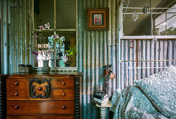 Antique chest of drawers next to metal bed in vintage bedroom with corrugated iron wall