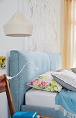 A light blue double bed with padded headboard and a chair with a clip lamp next to it