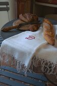 Besticktes, weißes Tuch auf Bistrotisch mit Brotmesser, Schneidebrett und Brot