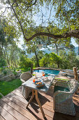 Rattan armchairs around set breakfast table on wooden terrace with swimming pool in background