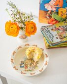 Blumensträußchen und angebissener Muffin auf Kinderteller vor buntem Kinderbuchstapel