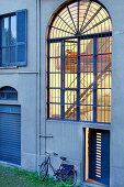 Bicycle leaning on exterior wall of house below large arched window