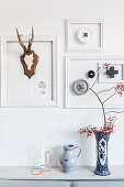 Arrangement of white picture frames and hunting trophies above two vases