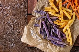 Variety of french fries traditional potatoes, purple potato, carrot served with salt on baking paper over brown texture background