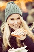 Winterlich gekleidete junge Frau isst eine Waffel auf Weihnachtsmarkt