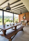 Long wooden dining table and bench in front of lounge area in interior with glass wall