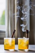 Two Golden Whiskey cocktails with burning cinnamon sticks and ice cubes
