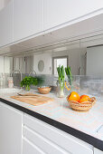 White kitchen counter with wooden chopping board, fruit bowl, vegetables and herbs on worksurface