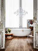 Freestanding bathtub in front of window, designer chandelier above, round table and metal chair in bathroom