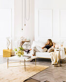 Young woman on white couch in front of white cassette paneling, in the foreground a coffee table with a bouquet of white flowers