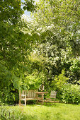 Sitzplatz in frühsommerlichem Garten mit Holzbank, Tisch und Klappstuhl