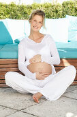 Schwangere Frau mit weißem Outfit im Lotussitz auf der Terrasse