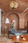 Coffee table, leather sofa and standard lamp in open-plan interior with brick walls
