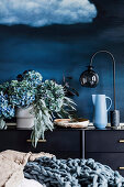 Vase with hydrangeas and eucalyptus branches, bowl, jug and table lamp on sideboard against blue wall