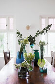 Glasvasen mit Blumen und Blätterzweigen auf rustikalem Esstisch