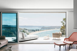 View through open sliding door to terrace with daybed, side table and club chair and to the sea