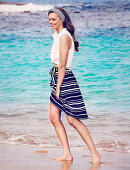 A young woman on a beach wearing a white, sleeveless blouse and a blue-and-white striped skirts