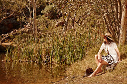 Junge Frau mit Hut in weißer Bluse und Rock am Flussufer sitzend