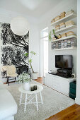 Runder, weißer Beistelltisch auf hellem Teppich, Fernsehschrank, Wandregale und zwei Stühle vor Wandposter im Wohnzimmer