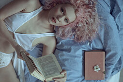 Junge Frau mit rosa Haaren in Unterwäsche liegt mit einem Buch auf dem Bett