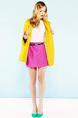 A blonde woman wearing a light blouse, a pink skirt and a yellow coat