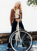 A blonde woman wearing a ruffled blouse, a brown jacket and jeans on a bike