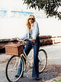 A blonde woman wearing sunglasses, a light blue jacket and jeans on a bike