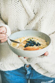 Woman in woolen sweater eating vegan almond milk oatmeal porridge with berries, fruit and almond