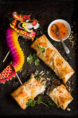 Two homemade vegetable spring rolls one torn open sprinkled with chilli flakes and sesame seeds and fresh coriander with sweet chilli sauce and a celebration paper dragon