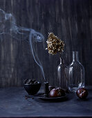 Still-life arrangement of dark fruit, glass bottles and candle smoke