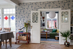 Open double doors painted white, vintage serving trolley and house plants against patterned wallpaper