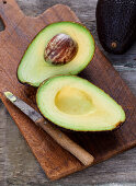 A halved avocado with a stone and a knife on a rustic wooden board