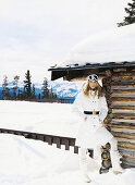 Blonde Frau in weißem Steppmantel und Boots an Holzhütte