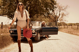 A brunette woman standing on a country road with a blonde woman in a car in the background