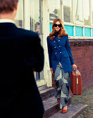 A brunette woman wearing a blue coat and a blue patterned trousers holding a suitcase