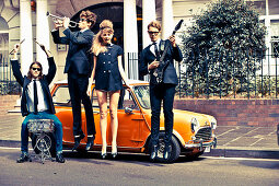 Elegantly dressed young people with musical instruments in front of a Mini