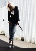 A young woman wearing a black blazer and rolled-up jeans standing in front of a corrugated wall