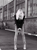 A blonde woman wearing a black jacket and jeans in front of an industrial building (black-and-white shot)