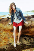 A young woman wearing a knitted jumper, a denim jacket and a red skirt on a beach