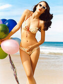A brunette woman on a beach wearing a bikini and holding balloons