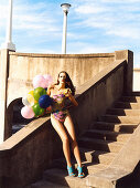 A brunette woman on a flight of steps wearing a bathing suit and holding balloons