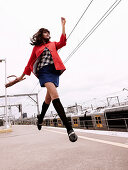 A brunette woman wearing a red coat, a blue skirt and black knee socks at a station