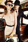 A young woman wearing sunglasses and a dress and young man wearing a hat and a black suit on a train