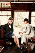 A young woman wearing a black suit and a young man wearing a hat and a light suit sitting on a train