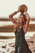 A blonde woman on a beach wearing a green dress holding an exotic fruit above her head