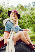 A blonde woman wearing a hat and a scarf, a denim blouse and a skirt sitting on a tractor