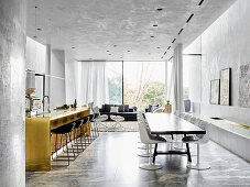 Long dining table with shell chairs, polished brass kitchen island with bar stools in an elegant, open living room with marble tiles