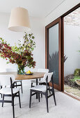 Round table with branches of leaves and white chairs in front of open sliding patio door