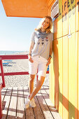 A blonde woman wearing a striped top and shorts leaning against a beach hut