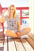 A blonde woman wearing a striped top and shorts sitting against a beach hut
